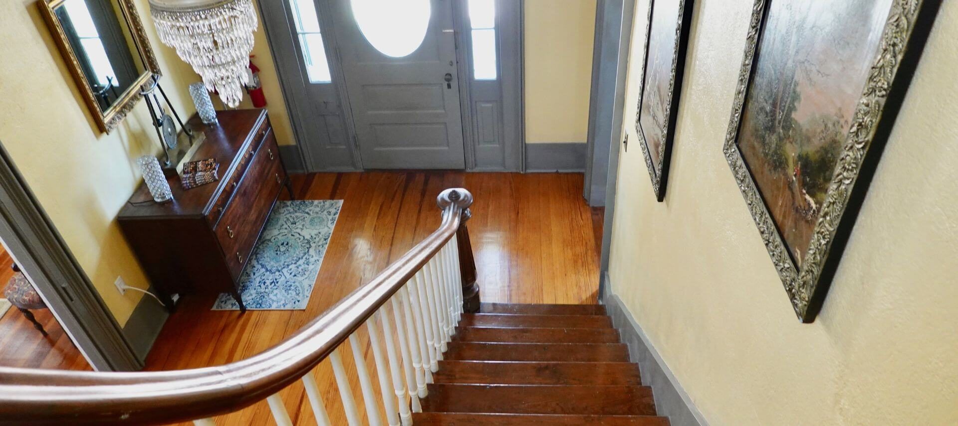 entryway with wood stars, white spindles and wood handrail, with wood floors, a grey door with an oval window and vertical side windows, cream-colored walls with grey trim