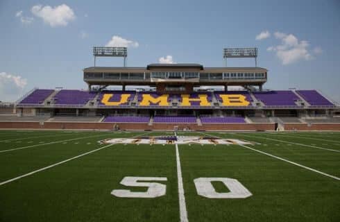 a football stadium with purple seats in the stand and the letters UMHB, along with green turf and a 50 yard line.