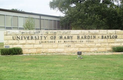 A cream colored brick wall with the words University of Mary Hardin-Baylor Chartered by Republic of Texas 1845, with a building with vertical windows behind it, green grass in front of it, and green trees and bushes