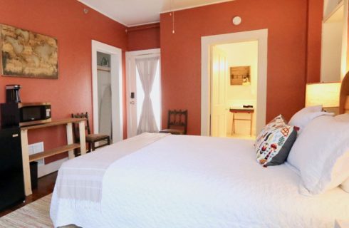 bedroom with salmon colored walls, a bed with white bedding and a colorful decorative pillow, along with a mini fridge, coffeemaker, and microwave. Also a closet with iron and ironing board, and a door opening to a private bathroom.