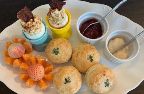 A white decorative oval plate with breakfast treats such as mini muffins, strawberries, orange slices, poached eggs, and mini crocks of butter and jam