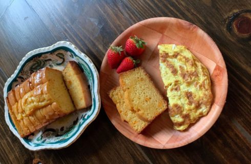 dark wood dining table with a green and white plate with sliced orange sweet bread and a wood plate with a yellow and omelet, red strawberries, and 2 pieces of sliced bread.