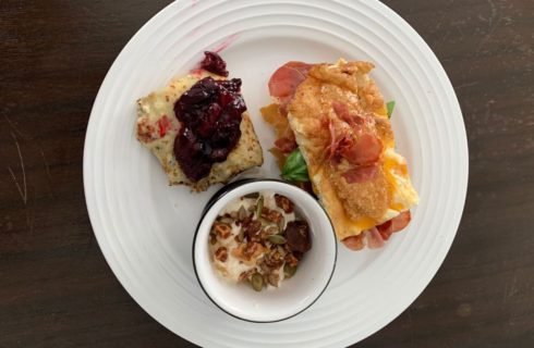 a white plate with breakfast foods of a ham and egg breakfat bake, a bowl of yogurt with granola, and a piece of bread with jam