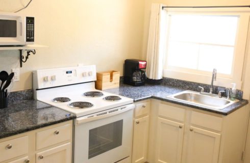 kitchen with cream walls and cupboards black granite countertops, white stove and oven, microwave and coffeemaker, metal sink, and cooking utensils