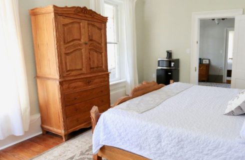 bedroom with light green walls, white trim, a bed with a wood footboard and white bedding, a wood armoire, and a small fridge with microwave and coffeemaker on top, along with windows and white curtains