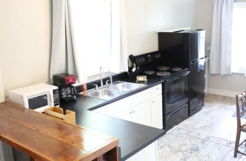 a kitchen with a black refrigerator and stove, white cupboards, metal sinks, coffeemaker microwave, table and chair, and wood breakfast bar