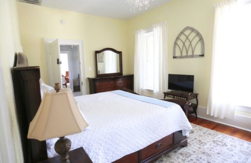 Bedroom with yellow walls with white trim, wood floors, tapestery area rug, a bed with whit bedding, a lamp on a nighstand, a dresser with mirror, a flat screen TV on a TV stand and windows with white curtains