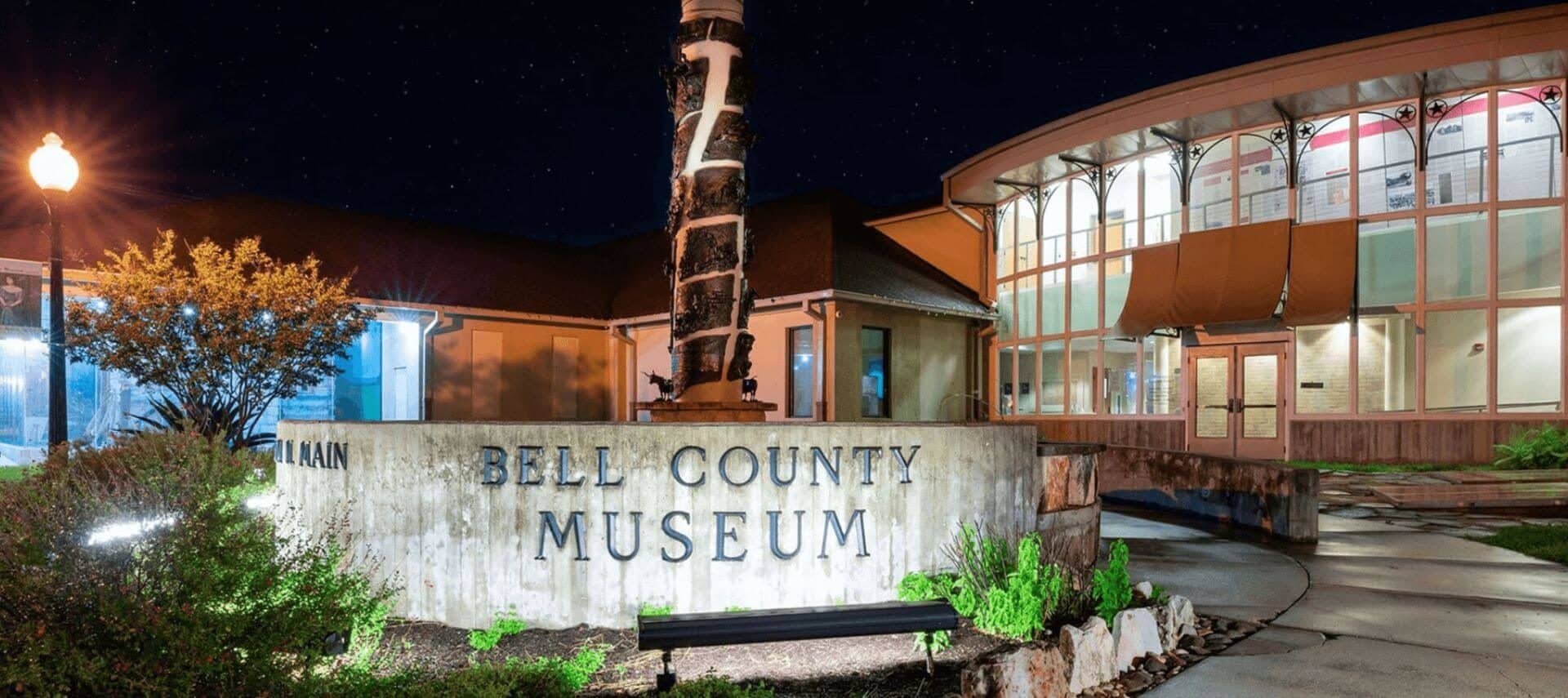 a night time photo of the Bell County Museum with multiple story glass windowed circular building and green bushes and srubs, along with a concrete walkway