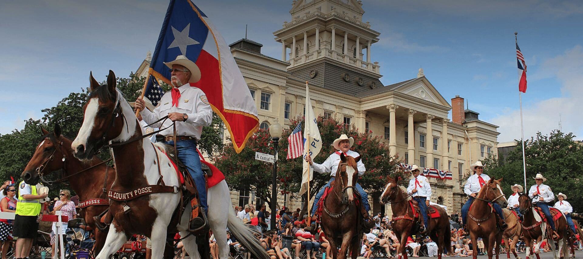 A group of men and women on horseback wearing white shirts, blue jeans, and white cowboy hats, holding various flags representing the US and Texas, in front of a multi story brick building