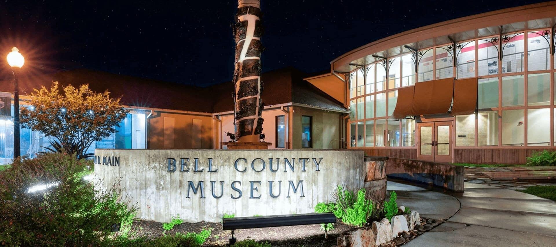 a night time photo of the Bell County Museum with multiple story glass windowed circular building and green bushes and srubs, along with a concrete walkway