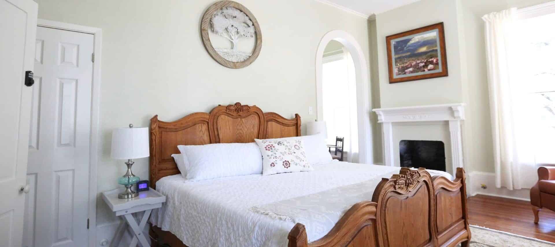 bedroom with light green walls, white doors and trim, wood floors, a bed with a carved wood headboard and footboards, white bedding, side tables with lamps, a fireplace with white mantle, and a carved circular oak tree on the wall